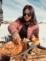 A person wearing a pink jacket and sunglasses is sitting in a snowy landscape, enjoying a meal. They are dipping bread into a large bowl of a tomato-based dish. Various other foods and drinks are spread out on the surface in front of them, including cups and fruit.