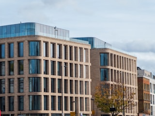 A modern office building with a brick facade, large glass windows, and multiple stories. The architecture features rectangular patterns and a rooftop with blue-tinted glass walls. In the foreground, a tree with autumn leaves and street signs are visible.