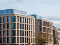 A modern office building with a brick facade, large glass windows, and multiple stories. The architecture features rectangular patterns and a rooftop with blue-tinted glass walls. In the foreground, a tree with autumn leaves and street signs are visible.