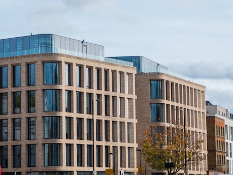 A modern office building with a brick facade, large glass windows, and multiple stories. The architecture features rectangular patterns and a rooftop with blue-tinted glass walls. In the foreground, a tree with autumn leaves and street signs are visible.
