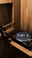 A wooden shelf unit with a vinyl record player placed on a wooden surface. A pair of headphones hangs on the side of the shelf. The record player has a unique slipmat with graphics and text on it.