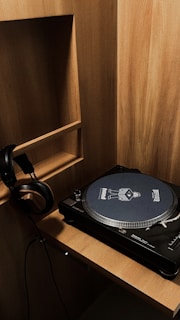 A wooden shelf unit with a vinyl record player placed on a wooden surface. A pair of headphones hangs on the side of the shelf. The record player has a unique slipmat with graphics and text on it.