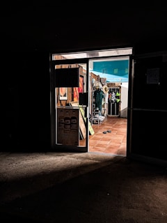 A dimly lit entrance to a store with warm, inviting light coming from inside. The store displays various items, including hats and clothing. The floor is tiled with terracotta tiles, and there are flip-flops on the ground near the entrance.