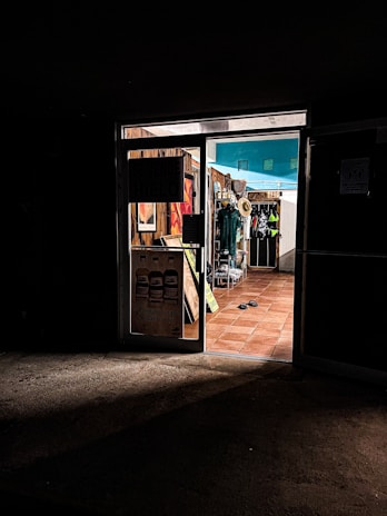 A dimly lit entrance to a store with warm, inviting light coming from inside. The store displays various items, including hats and clothing. The floor is tiled with terracotta tiles, and there are flip-flops on the ground near the entrance.