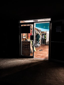 A dimly lit entrance to a store with warm, inviting light coming from inside. The store displays various items, including hats and clothing. The floor is tiled with terracotta tiles, and there are flip-flops on the ground near the entrance.