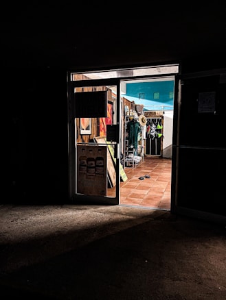 A dimly lit entrance to a store with warm, inviting light coming from inside. The store displays various items, including hats and clothing. The floor is tiled with terracotta tiles, and there are flip-flops on the ground near the entrance.