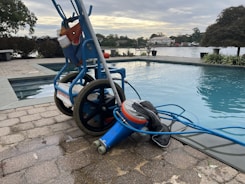 A wheeled cart with a pool cleaning device is parked next to an outdoor swimming pool. The cart holds various cleaning supplies and hoses. The pool is surrounded by a stone-tiled deck and overlooks a body of water with trees and houses in the background.