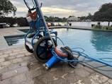 A wheeled cart with a pool cleaning device is parked next to an outdoor swimming pool. The cart holds various cleaning supplies and hoses. The pool is surrounded by a stone-tiled deck and overlooks a body of water with trees and houses in the background.