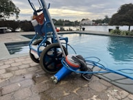 A wheeled cart with a pool cleaning device is parked next to an outdoor swimming pool. The cart holds various cleaning supplies and hoses. The pool is surrounded by a stone-tiled deck and overlooks a body of water with trees and houses in the background.