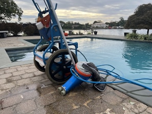 A wheeled cart with a pool cleaning device is parked next to an outdoor swimming pool. The cart holds various cleaning supplies and hoses. The pool is surrounded by a stone-tiled deck and overlooks a body of water with trees and houses in the background.