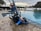 A wheeled cart with a pool cleaning device is parked next to an outdoor swimming pool. The cart holds various cleaning supplies and hoses. The pool is surrounded by a stone-tiled deck and overlooks a body of water with trees and houses in the background.