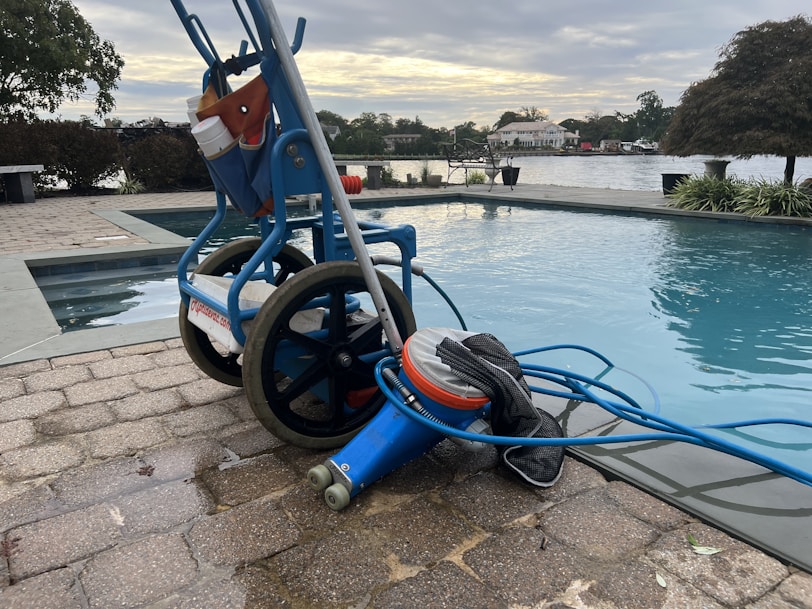 A wheeled cart with a pool cleaning device is parked next to an outdoor swimming pool. The cart holds various cleaning supplies and hoses. The pool is surrounded by a stone-tiled deck and overlooks a body of water with trees and houses in the background.