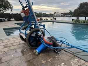 A wheeled cart with a pool cleaning device is parked next to an outdoor swimming pool. The cart holds various cleaning supplies and hoses. The pool is surrounded by a stone-tiled deck and overlooks a body of water with trees and houses in the background.