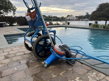 A wheeled cart with a pool cleaning device is parked next to an outdoor swimming pool. The cart holds various cleaning supplies and hoses. The pool is surrounded by a stone-tiled deck and overlooks a body of water with trees and houses in the background.