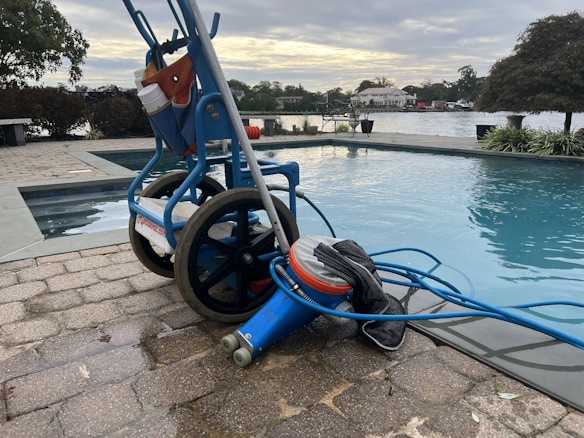 A wheeled cart with a pool cleaning device is parked next to an outdoor swimming pool. The cart holds various cleaning supplies and hoses. The pool is surrounded by a stone-tiled deck and overlooks a body of water with trees and houses in the background.