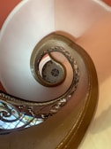 A spiral staircase with an ornate railing. The view is from above, focusing on the elegant curves and geometric patterns created by the stairs. The steps are bordered by a smooth, brown handrail, and the staircase descends into a circular mosaic design on the floor. Soft lighting enhances the warm, earthy tones, and stained glass is visible at the base.