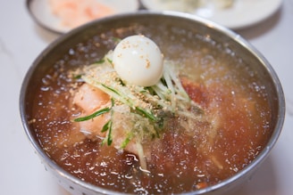 A bowl of Korean cold noodles, known as naengmyeon, is topped with slices of cucumber, sesame seeds, and a half-boiled egg, with a few slices of radish. The broth appears icy and refreshing, possibly made with beef or dongchimi.