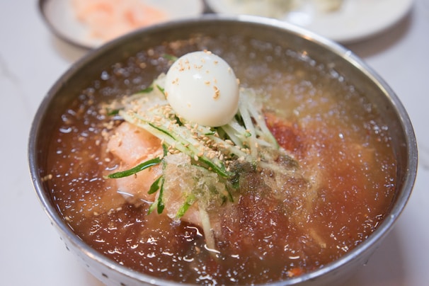 A bowl of Korean cold noodles, known as naengmyeon, is topped with slices of cucumber, sesame seeds, and a half-boiled egg, with a few slices of radish. The broth appears icy and refreshing, possibly made with beef or dongchimi.