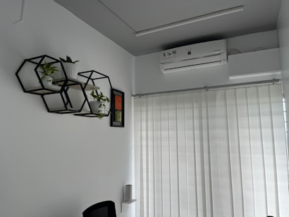 A modern office room with hexagonal wall shelves holding small potted plants and a framed picture. An air conditioning unit is mounted on the ceiling above vertical blinds covering a large window.