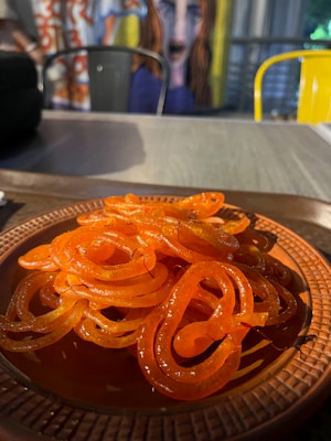 A plate of glossy, orange jalebis, an Indian sweet made from deep-fried batter soaked in sugar syrup, is placed on a textured brown plate. In the background, there is a mural featuring an abstract face with exaggerated features and vibrant colors. Yellow and metal chairs are partially visible.