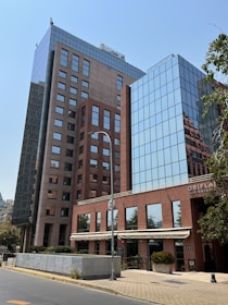 A modern multi-story office building with a mix of glass and brick façades. The structure's large reflective windows mirror the clear blue sky, while the lower brick sections provide a contrast. A sidewalk runs alongside, with trees and potted plants adding greenery. A lamppost and street signs are present in front of the building.