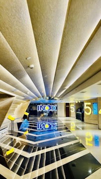 A modern and sleek cinema lobby features geometric patterns with reflective black and white flooring. The ceiling has angular design elements with recessed lighting, contributing to a futuristic aesthetic. Bright yellow elements, including the number '5' and signage, add a pop of color against the neutral backdrop. The word 'INOX' is prominently displayed in blue and yellow at the end of the hallway, with reflections creating a symmetrical visual effect.