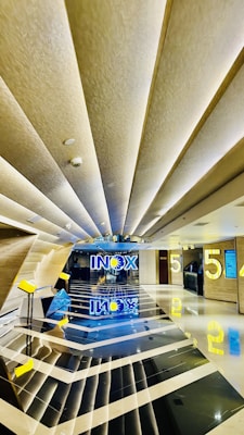 A modern and sleek cinema lobby features geometric patterns with reflective black and white flooring. The ceiling has angular design elements with recessed lighting, contributing to a futuristic aesthetic. Bright yellow elements, including the number '5' and signage, add a pop of color against the neutral backdrop. The word 'INOX' is prominently displayed in blue and yellow at the end of the hallway, with reflections creating a symmetrical visual effect.
