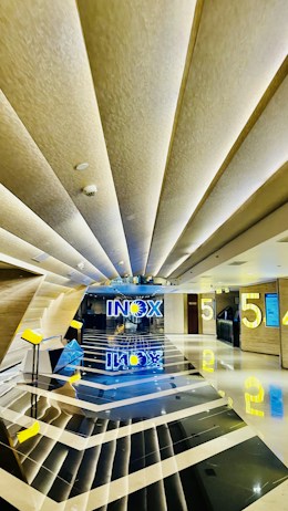 A modern and sleek cinema lobby features geometric patterns with reflective black and white flooring. The ceiling has angular design elements with recessed lighting, contributing to a futuristic aesthetic. Bright yellow elements, including the number '5' and signage, add a pop of color against the neutral backdrop. The word 'INOX' is prominently displayed in blue and yellow at the end of the hallway, with reflections creating a symmetrical visual effect.