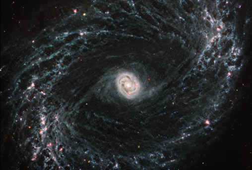 A stunning spiral galaxy with a bright central core surrounded by swirling arms of stars, dust, and gas. The arms are dotted with clusters of bright red and blue stars, creating a striking pattern against the dark backdrop of space.
