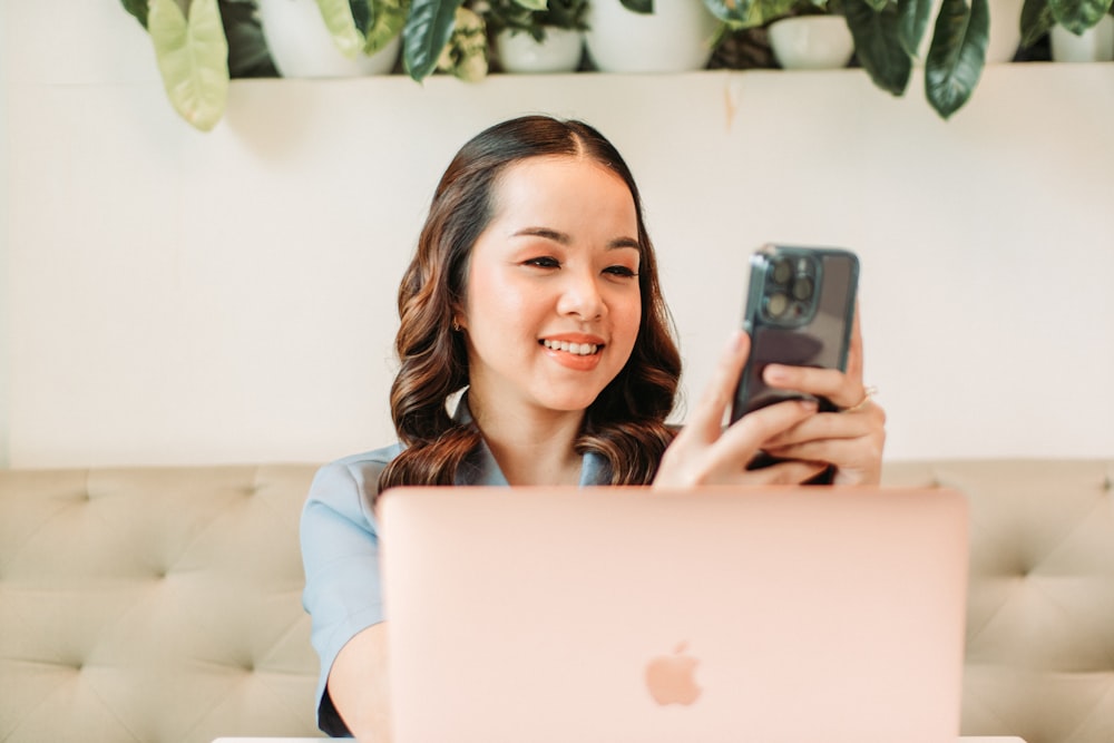 a woman sitting in front of a laptop holding a cell phone