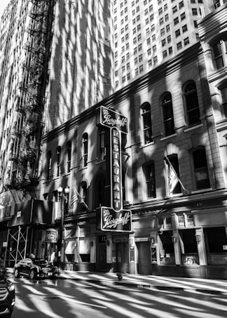 A black and white photograph of a city street showcasing a historic restaurant with a vintage sign. Tall buildings surround the area, casting intricate shadows and creating a dramatic play of light and darkness.