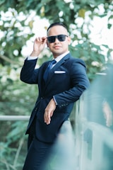 A man wearing a formal suit with a tie and pocket square leans against a glass railing while adjusting his sunglasses. The background is filled with lush green foliage, suggesting an outdoor setting.