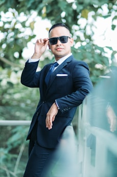 A man wearing a formal suit with a tie and pocket square leans against a glass railing while adjusting his sunglasses. The background is filled with lush green foliage, suggesting an outdoor setting.