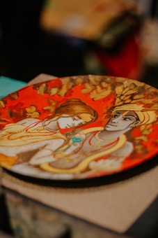 A decorative plate featuring a detailed painting of two figures with intricate clothing, set against a vibrant red and gold background. The figures exhibit classic artistic rendering with stylized expressions and flowing garments typical of traditional art styles.
