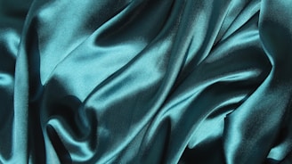 Smooth, flowing fabric with a silky texture and rich teal color, creating a luxurious appearance with light reflecting off the folds.