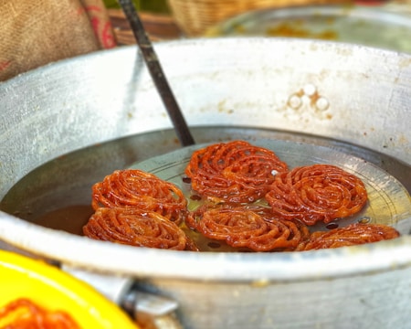 A metal pot containing several pieces of fried, orange-brown spiral-shaped snacks submerged in oil, possibly jalebi. A flat metal strainer is partially visible, used for lifting the snacks out of the oil.