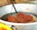 A metal pot containing several pieces of fried, orange-brown spiral-shaped snacks submerged in oil, possibly jalebi. A flat metal strainer is partially visible, used for lifting the snacks out of the oil.