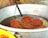 A metal pot containing several pieces of fried, orange-brown spiral-shaped snacks submerged in oil, possibly jalebi. A flat metal strainer is partially visible, used for lifting the snacks out of the oil.