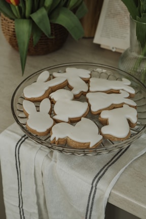 A glass plate is arranged with several cookies shaped like bunnies, chicks, and other Easter-themed figures, all covered in white icing. The plate sits on a white cloth with black stripes, placed on a countertop. In the background, there's a basket with green foliage and a partially visible open book.