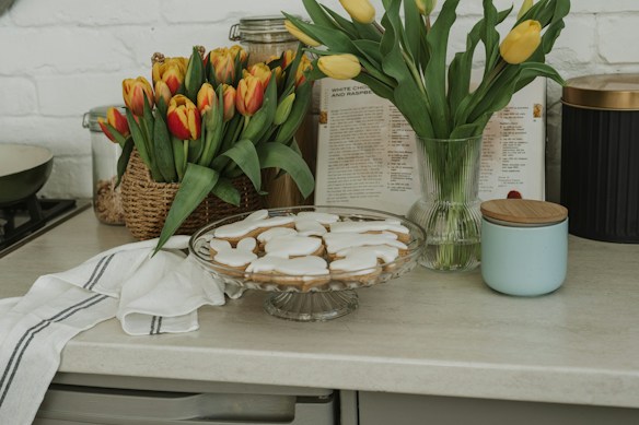 A kitchen counter is adorned with bouquets of tulips in vibrant orange and yellow. A glass cake stand holds iced cookies shaped like bunnies. A white towel with black stripes is draped on the counter next to a ceramic jar with a wooden lid. Behind the arrangement, a cookbook is open, adding a warm, inviting atmosphere.