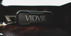 A rectangular, rugged Bluetooth speaker with the brand name 'VIDVIE' prominently displayed on the front sits on a wooden surface. The lighting is dim, casting shadows around the speaker, and a paper stapler is partially visible to the side.