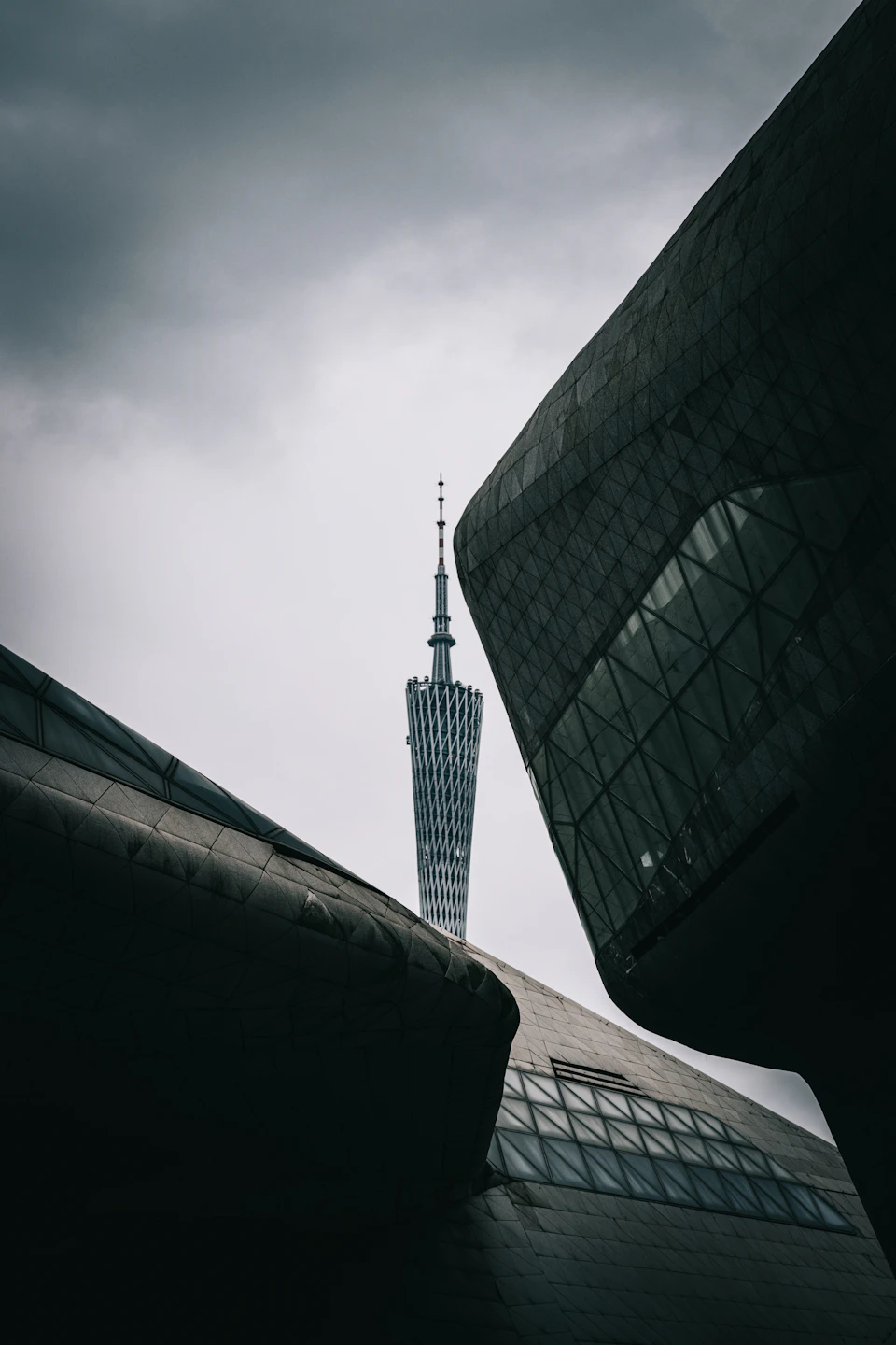 The image depicts a modern architectural structure with a unique triangular shape, featuring a glass facade that reflects the surrounding environment. The building is situated on a street corner and stands out against an overcast sky. In the background, there are two other buildings visible, one of which has a spire-like design at its peak.