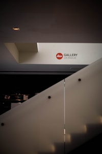 A modern architectural interior with a stairway leading to an upper level. The wall is adorned with a sign that reads 'Leica Gallery Los Angeles'. The lighting is minimal, casting soft shadows.