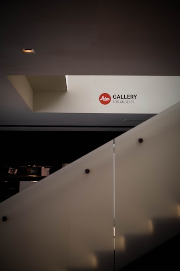 A modern architectural interior with a stairway leading to an upper level. The wall is adorned with a sign that reads 'Leica Gallery Los Angeles'. The lighting is minimal, casting soft shadows.