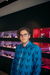 A person with medium-length hair and glasses wearing a blue shirt with a black V pattern. They are standing indoors with a background featuring display shelves and a red section with assorted items.