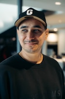 A person with a slight smile wearing a black cap with a letter 'G' on it and a black sweatshirt. The background is a softly focused indoor setting with a modern design.