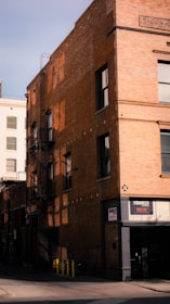 A multi-story brick building with large windows, featuring a fire escape ladder on its side. The building casts a shadow on the adjacent street. Signage for 'The Bunker' is visible near the entrance.