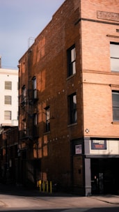 A multi-story brick building with large windows, featuring a fire escape ladder on its side. The building casts a shadow on the adjacent street. Signage for 'The Bunker' is visible near the entrance.