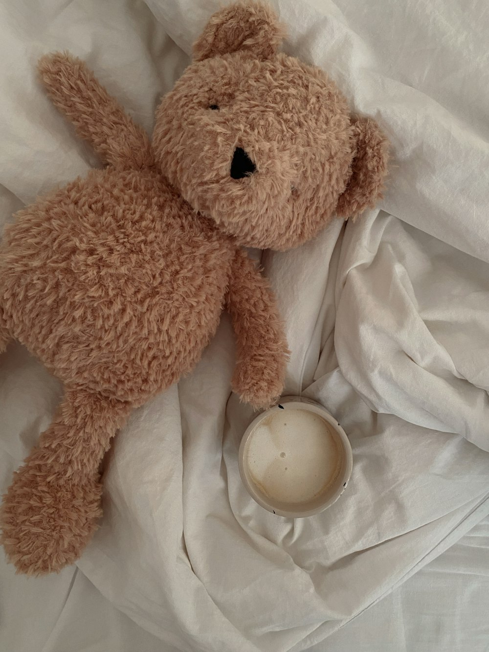 a brown teddy bear laying on top of a bed next to a cup