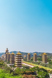 A series of traditional pagodas with golden rooftops are set against a backdrop of rolling green hills. A large Buddha statue is visible at the end of a pathway lined with greenery and landscaping.